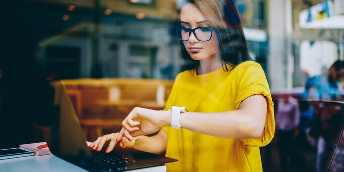 Young businesswoman in eyeglasses looking at display of wristwatch and managing time while checking email on laptop