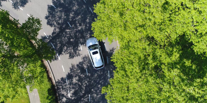 Aerial following car top-down view this grey colored station wagon is driving over two way street corner green trees on both sides of street