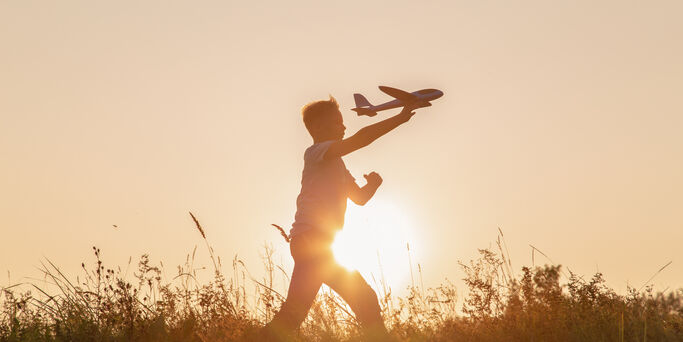 Black silhouette of cute happy cheerful child running fastly along grassy hill at countryside holding big toy plane in hand